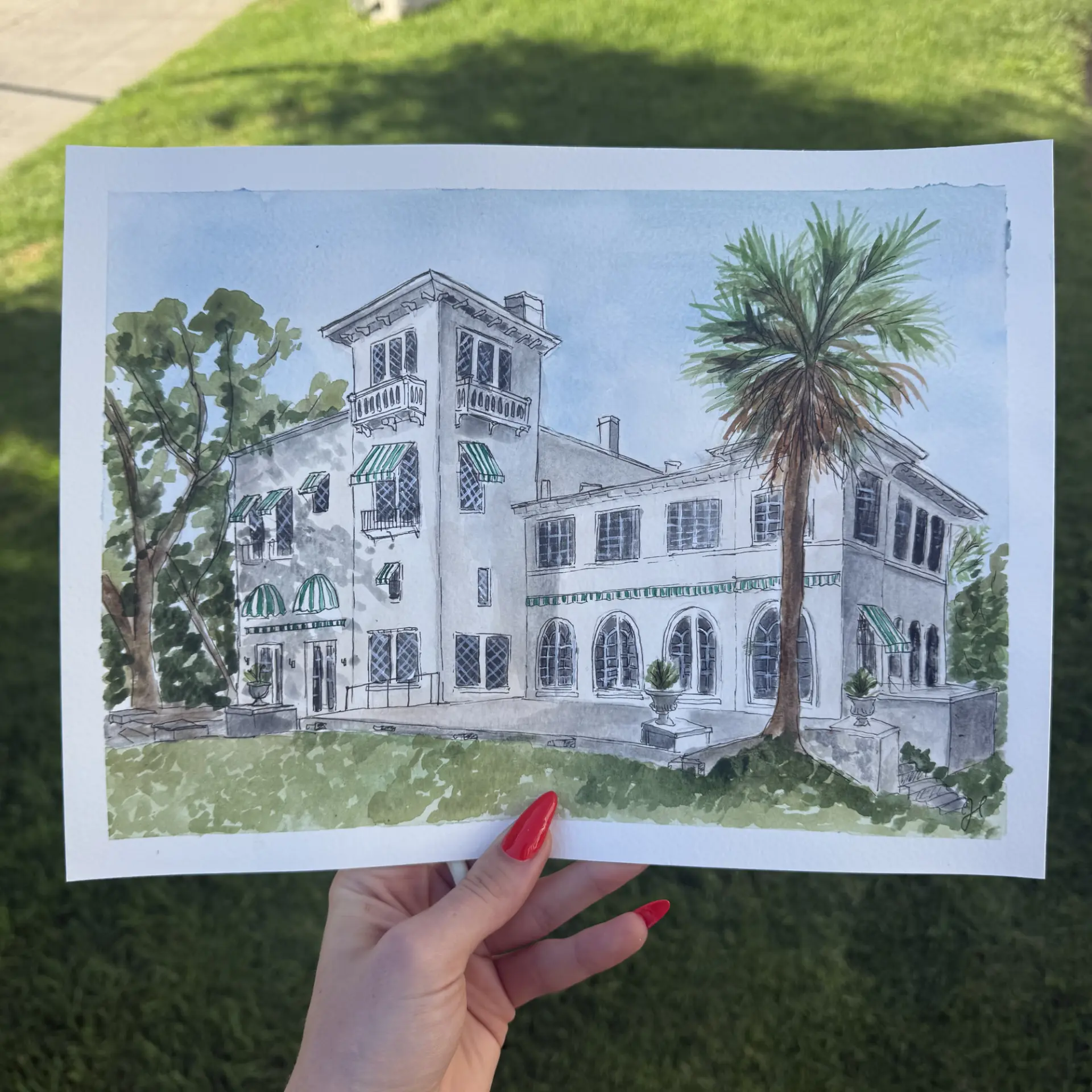 Hand holding a watercolor painting of a Mediterranean-style mansion